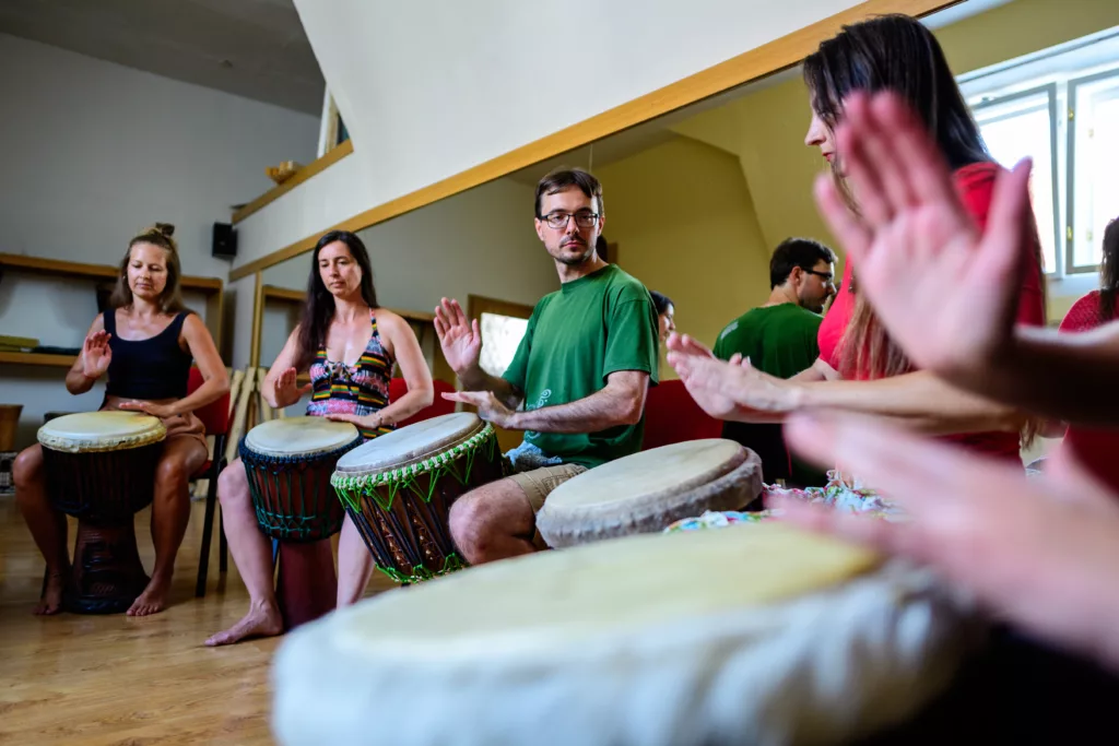 Tomáš Oplatek vede lekci bubnování: pět účastníků včetně Tomáše v zeleném tričku hraje na djembe bubny v kruhu. Dvě ženy vlevo, další účastníci vpravo. Lekce probíhá v interiéru s dřevěnou podlahou a přirozeným světlem.